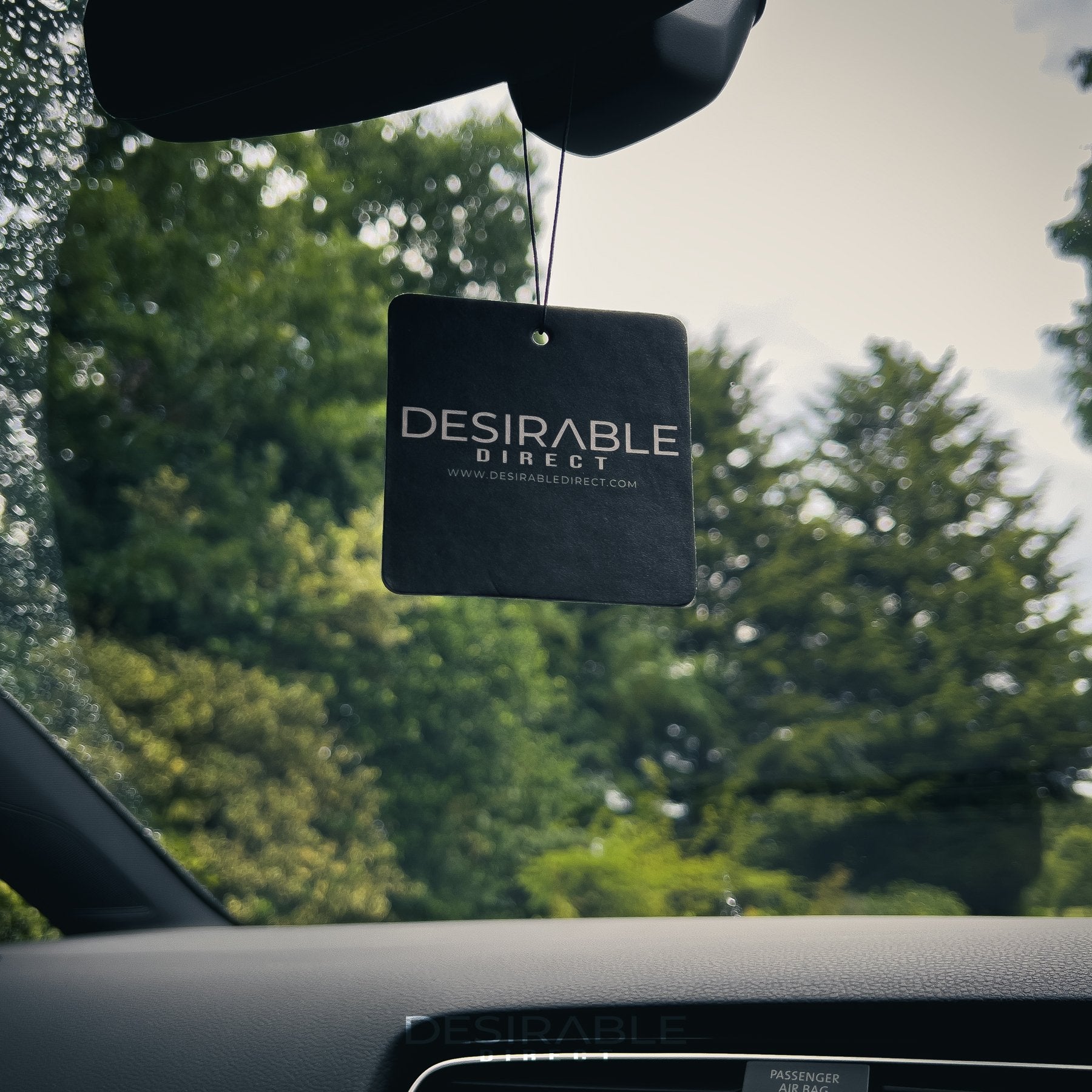 Car care black car air freshener with desirable direct and desirabledirect.com printed on the front, hanging in the front of a volkswagen golf r with trees in the background. This image showcases the air freshener as a bundle.