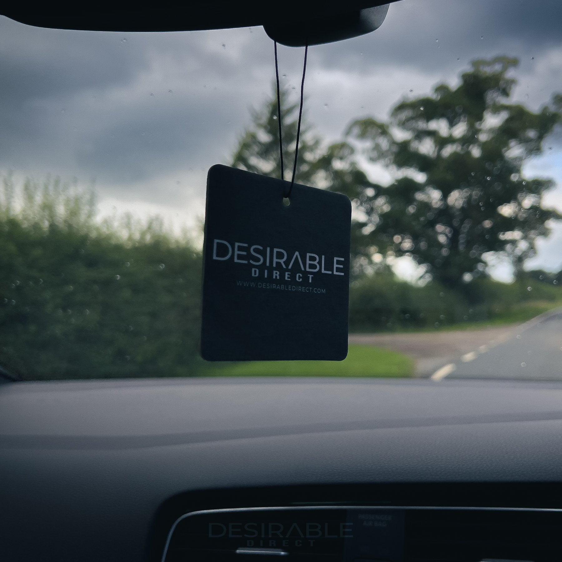 Car care black car air freshener with desirable direct and desirabledirect.com printed on the front, hanging in the front of a volkswagen golf r while driving on the road. This image showcases the air freshener as a bundle.