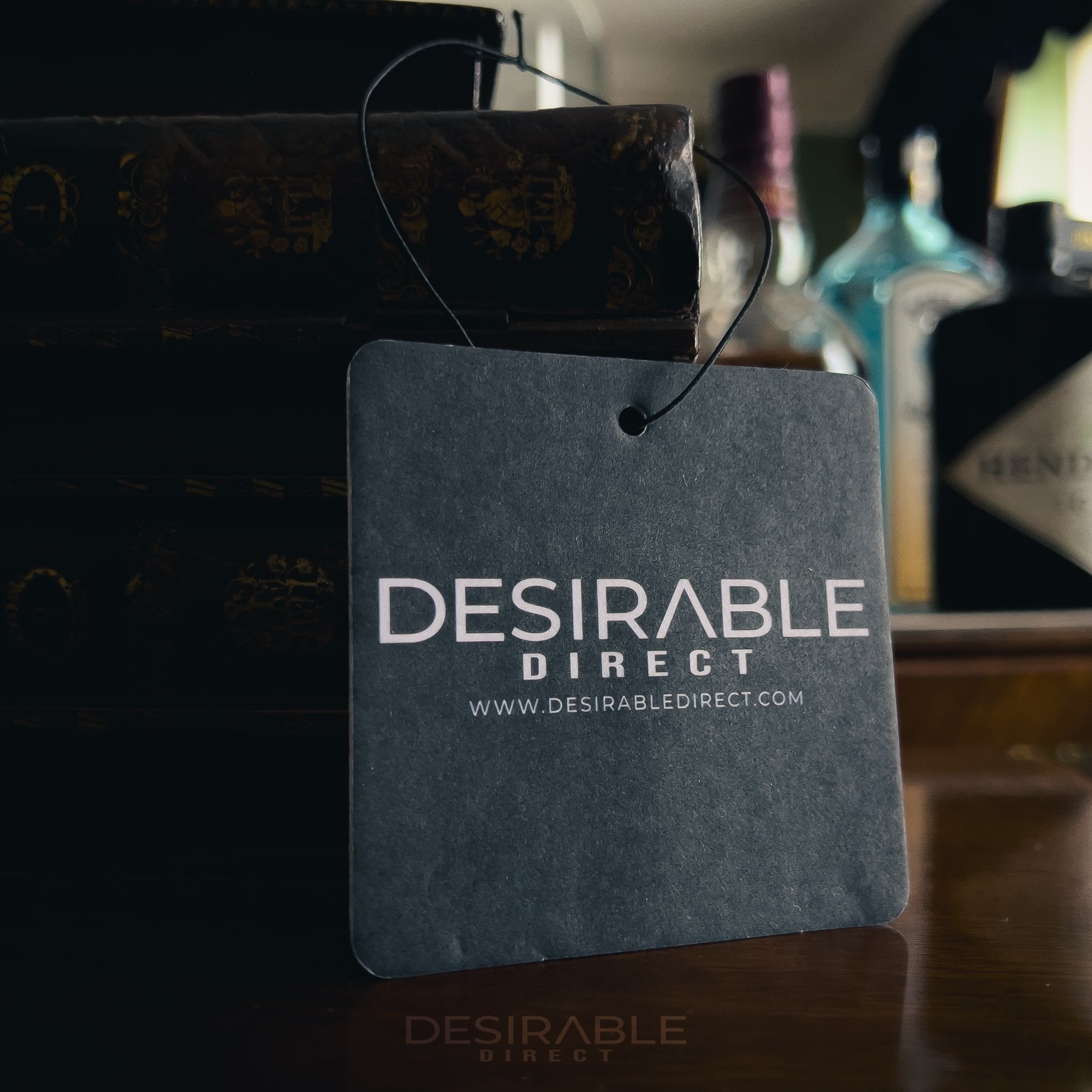 Car care black car air freshener with desirable direct and desirabledirect.com printed on the front, displayed on a brown table in a room with bottles in the background.