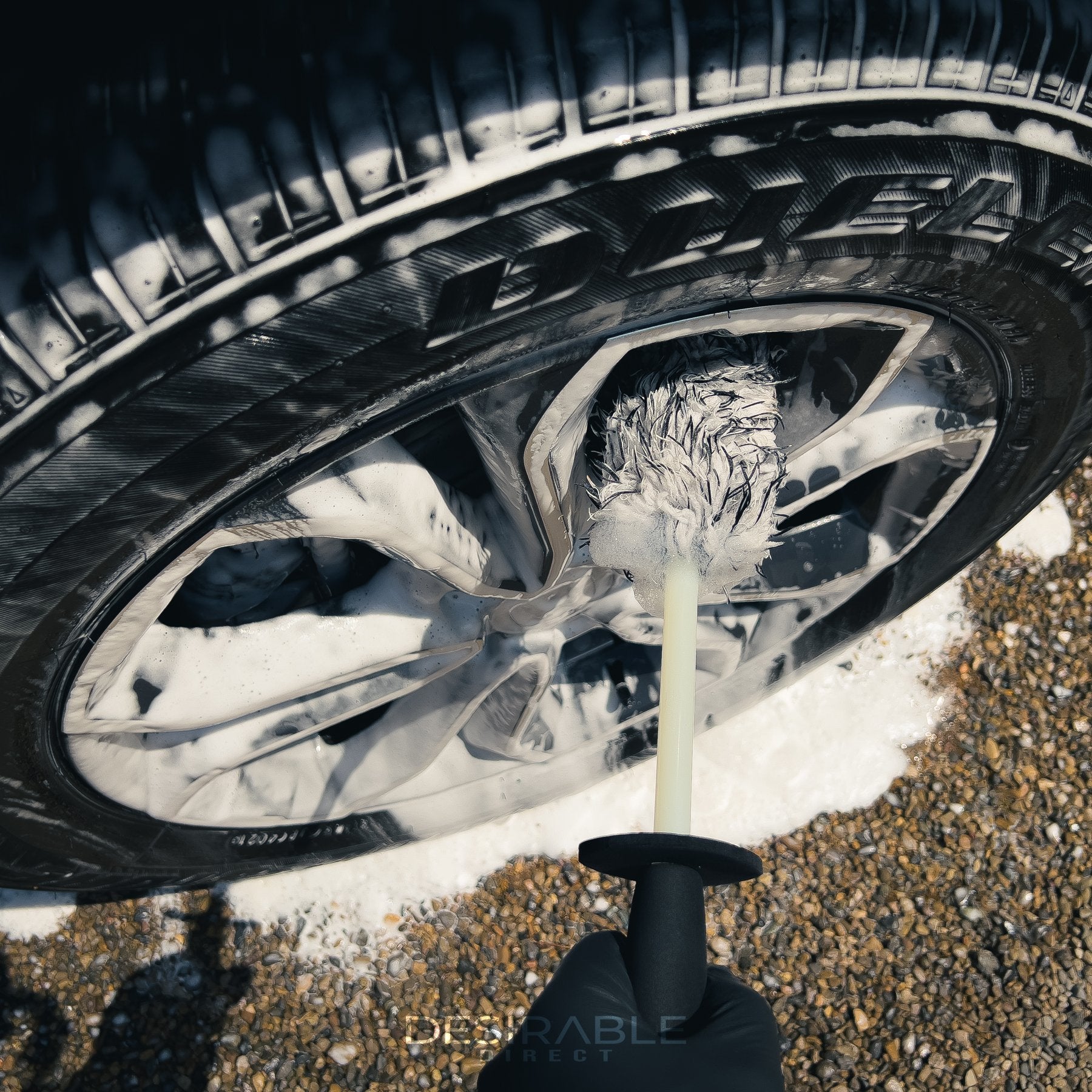 Car care microfibre black and white alloy wheel brush cleaning the alloy wheels of a grey car. The wheel is covered in car shampoo.