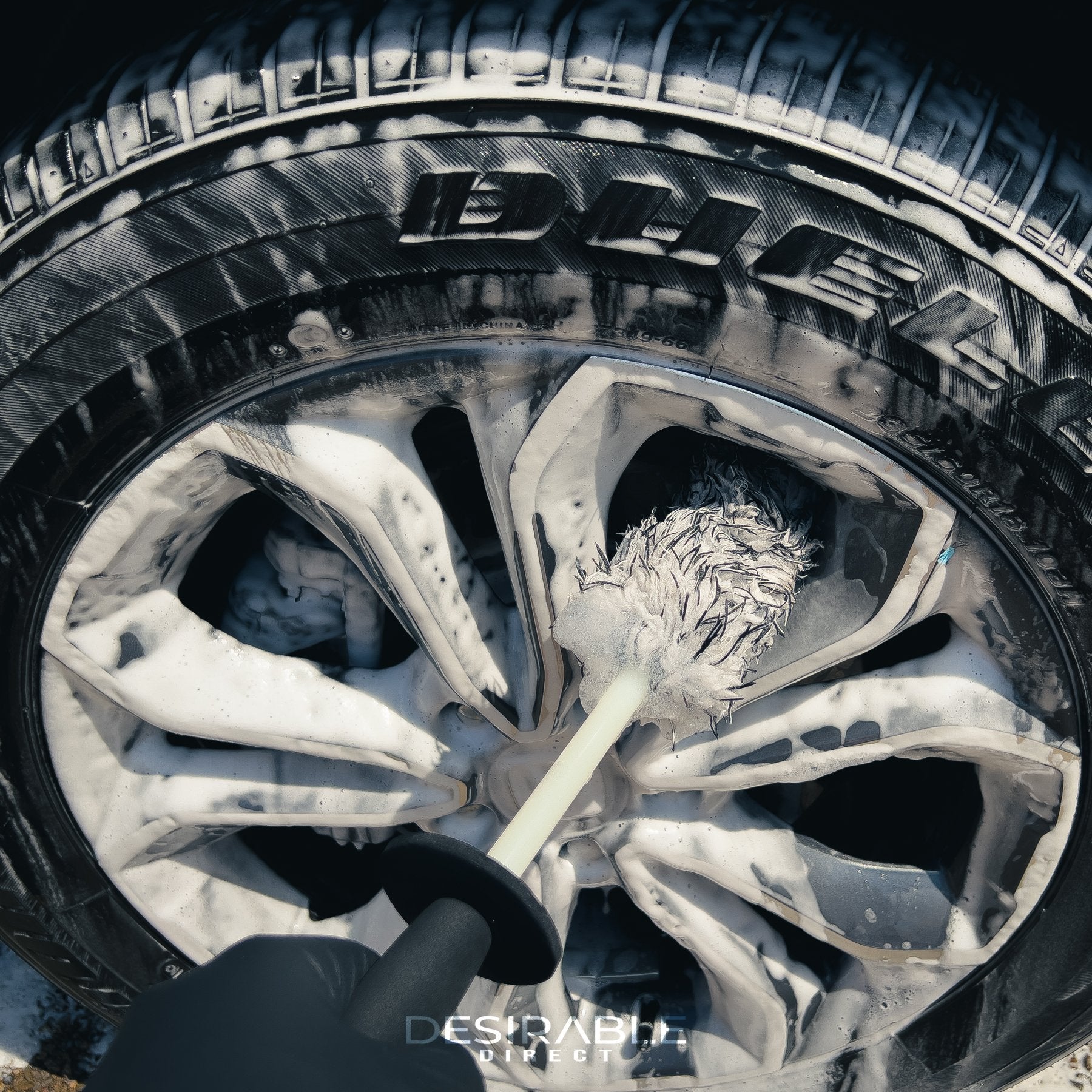 Car care microfibre black and white alloy wheel brush cleaning a silver and grey alloy wheel. The wheel is covered in car shampoo. The person holding the brush is wearing black gloves.