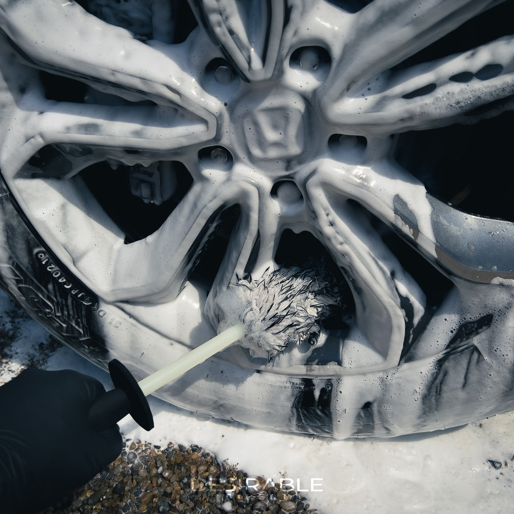 Car care microfibre black and white alloy wheel brush cleaning the bottom of a silver and grey alloy wheel. The wheel is covered in car shampoo. The person holding the brush is wearing black gloves.