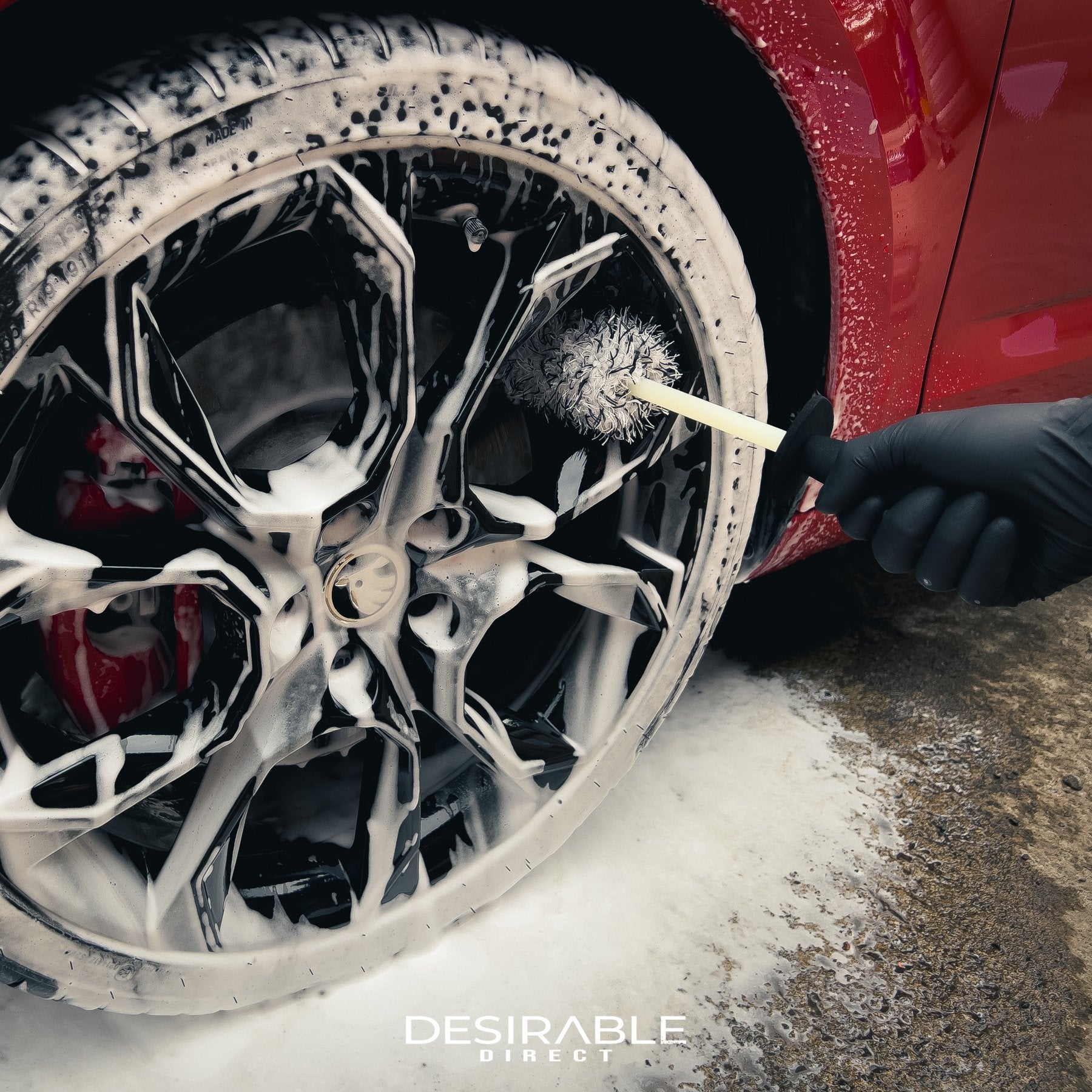 Car care microfibre black and white alloy wheel brush cleaning a black alloy wheel on a red car. The wheel is covered in car shampoo. The person holding the brush is wearing a black glove.