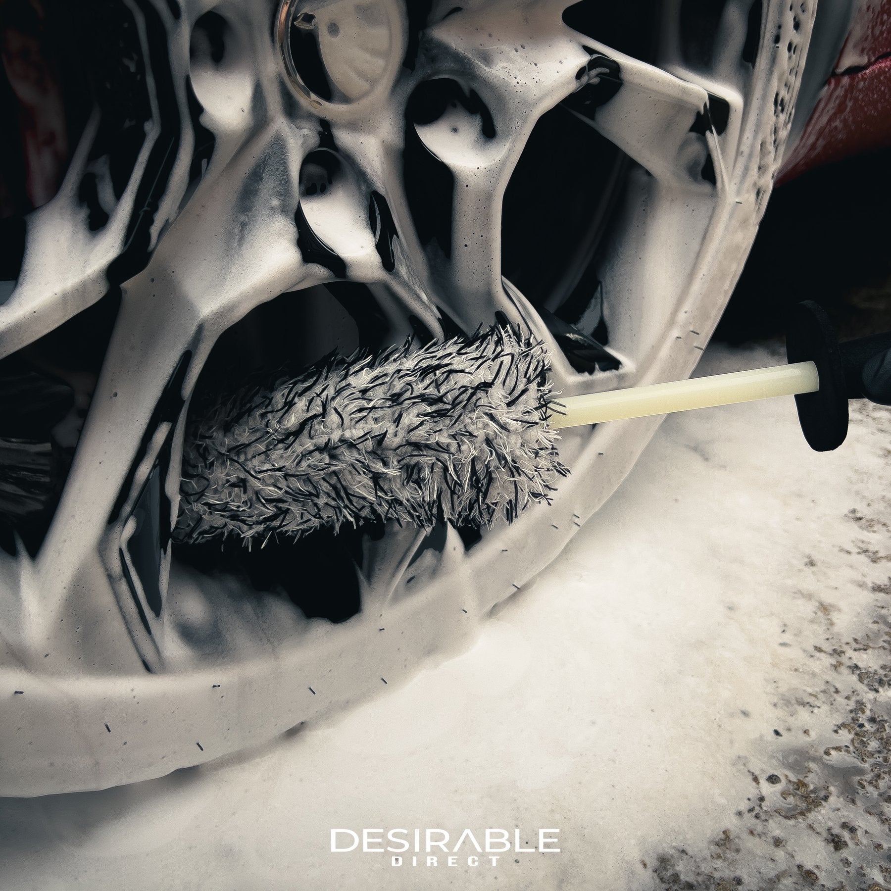 Car care microfibre black and white alloy wheel brush cleaning the bottom of a black alloy wheel on a red car. The wheel is covered in car shampoo.