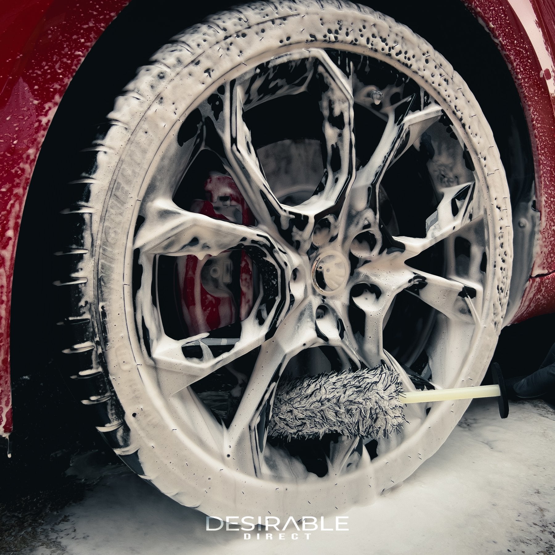 Car care microfibre black and white alloy wheel brush cleaning the bottom section of a black alloy wheel on a red car. The wheel is covered in car shampoo.