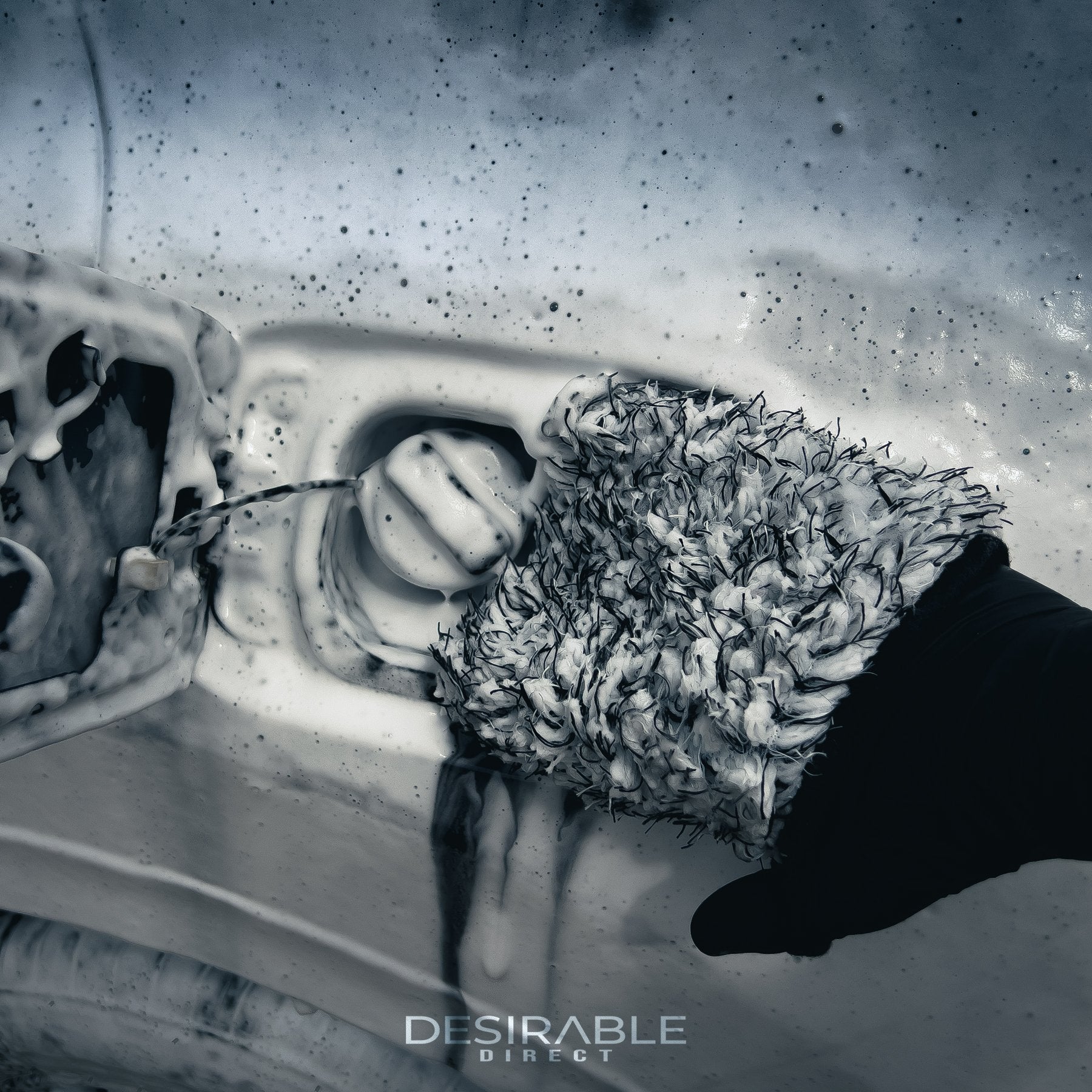 Car care microfibre black and white wheel and trim wash mitt cleaning the fuel filler cap of a grey car covered in car shampoo. The person holding the mitt is wearing black gloves.