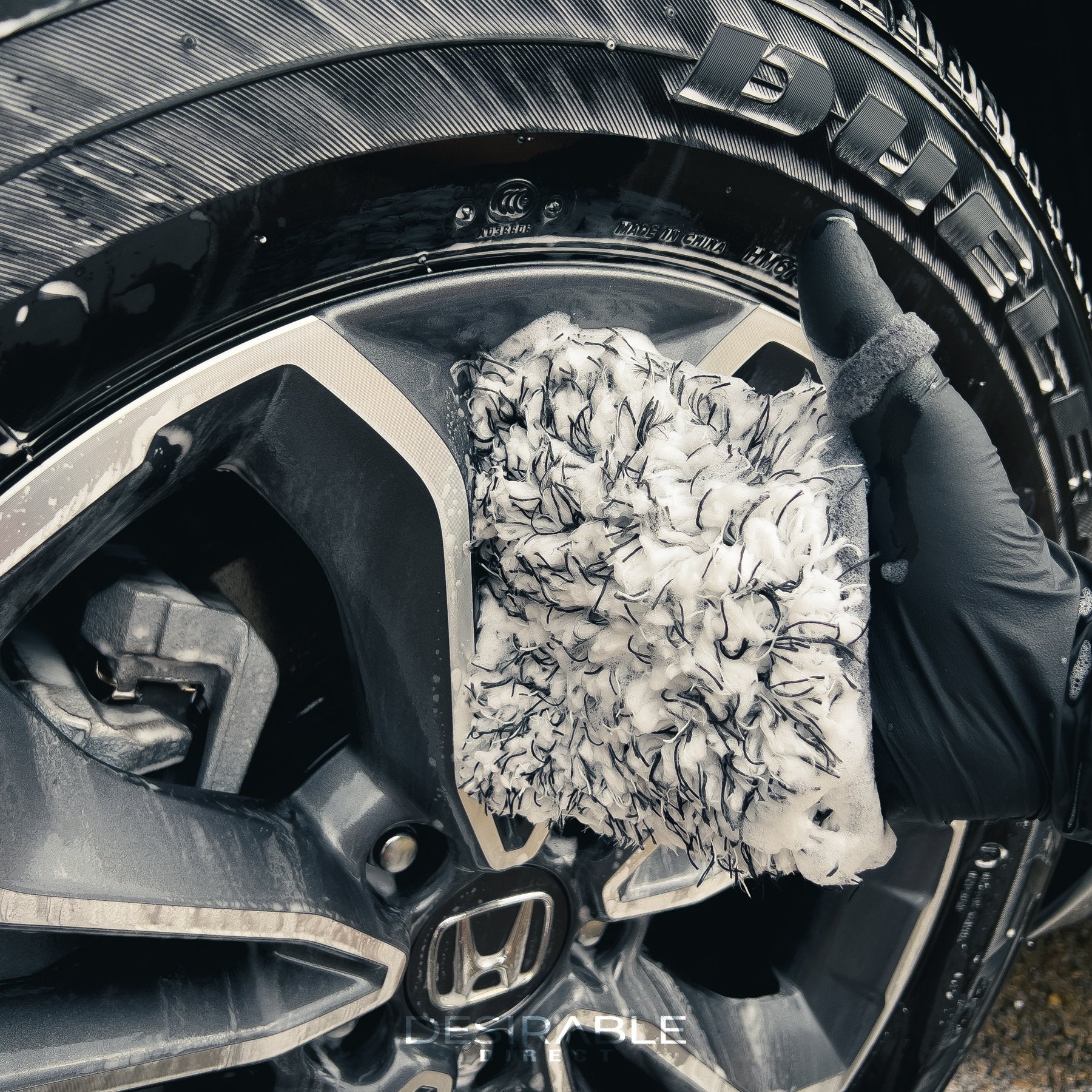 Car care microfibre black and white wheel and trim wash mitt cleaning the silver alloys of a grey car covered in car shampoo. The person holding the mitt is wearing black gloves.