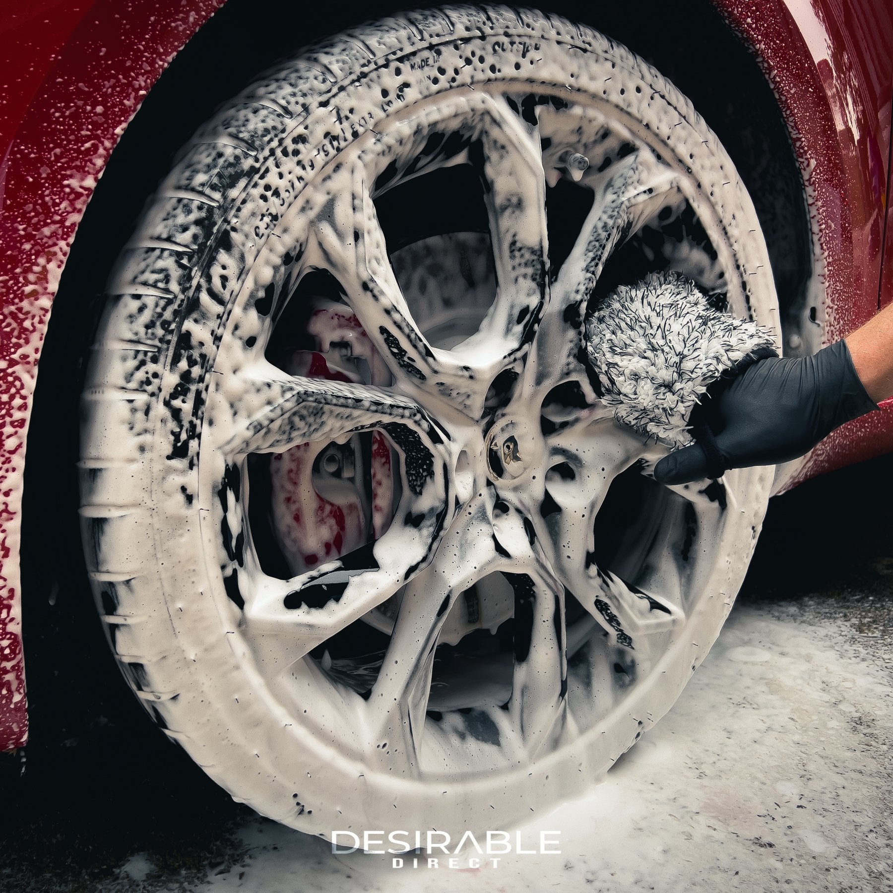 Car care microfibre black and white wheel and trim wash mitt cleaning the wheels of a red car with black wheels. The wheels are covered in car shampoo. This photo is from a different angle to showcase the product.