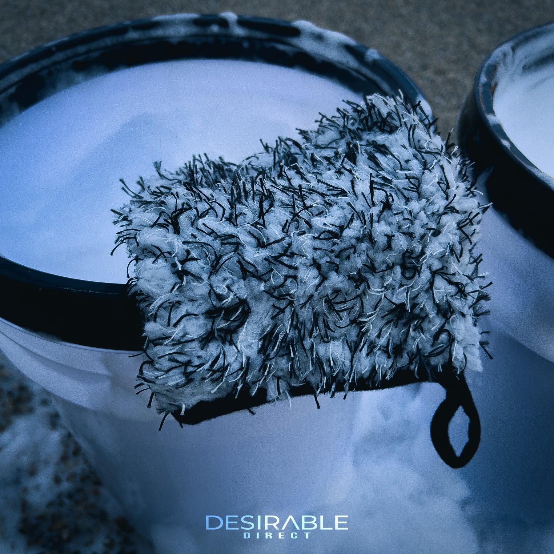 Car care microfibre black and white wheel and trim wash mitt displayed on the side of a car cleaning wash bucket filled with water and car shampoo.