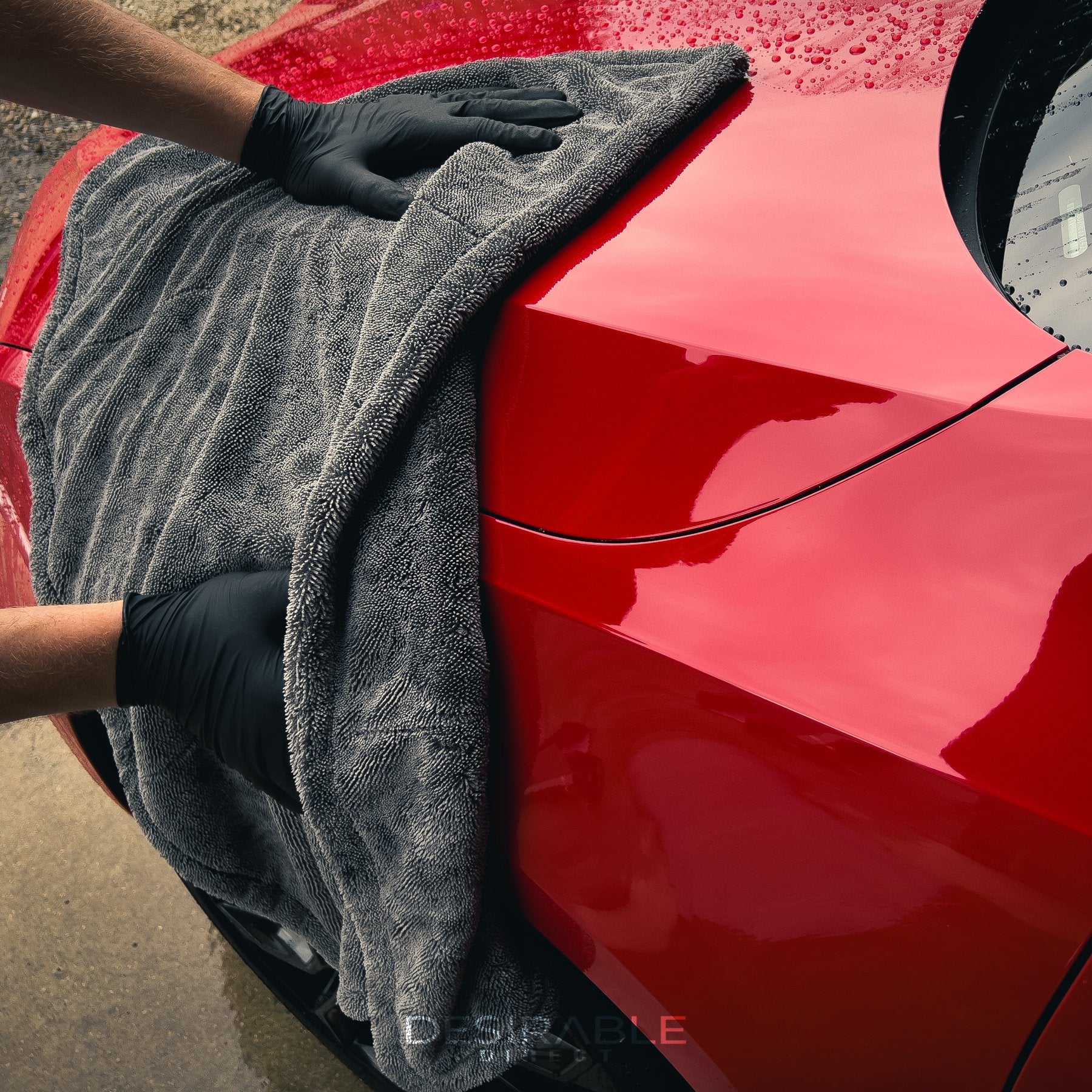 Car care large twisted pile grey drying towel drying a wet red car. Somebody is holding the towel while drying the front of the car wearing black gloves. The photo shows the drying performance as you can see the water has gone.
