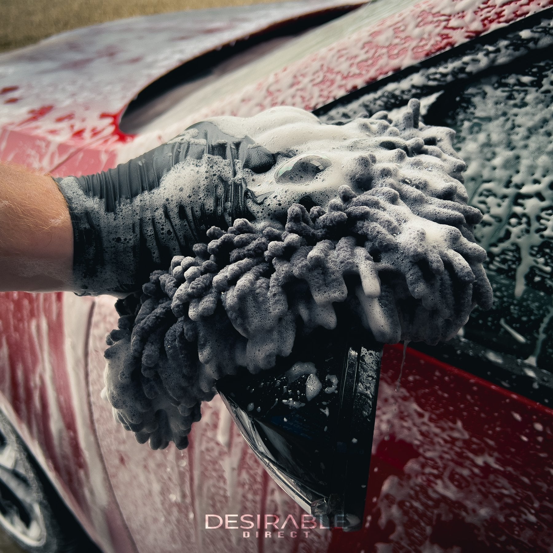Car care large grey noodle wash pad. The wash pad is covered in car shampoo, with a hand wearing a black glove moving the pad across the paintwork of the passenger side wing mirror of a red car in the cleaning process.