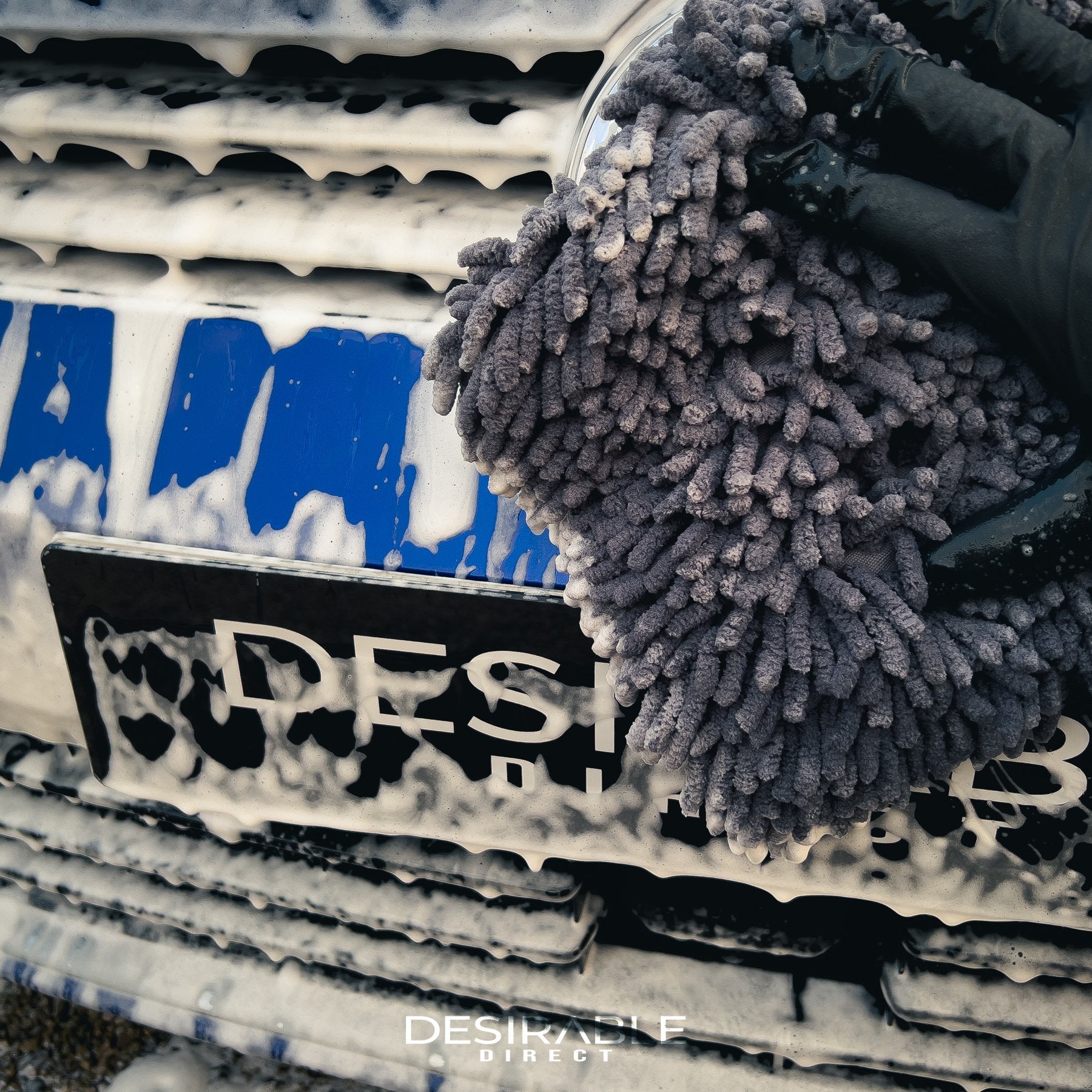 Car care large grey noodle wash pad. The wash pad is covered in car shampoo, with a hand wearing a black glove moving the pad across the paintwork of the front of a blue car.