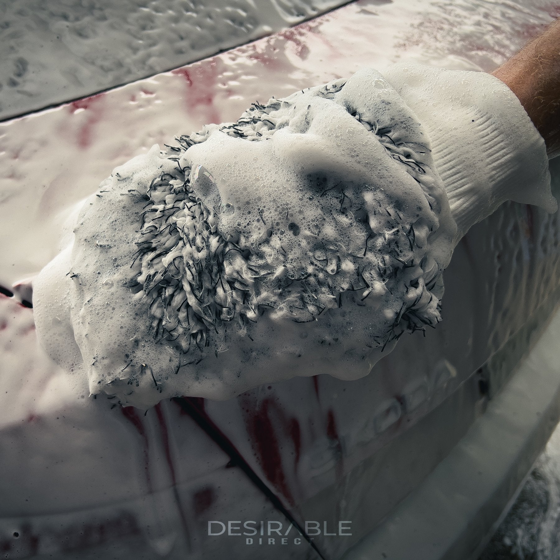 Car care microfibre black and white cleaning wash mitt cleaning the back of a red car covered in car shampoo. Somebody is holding the mitt while cleaning the car.