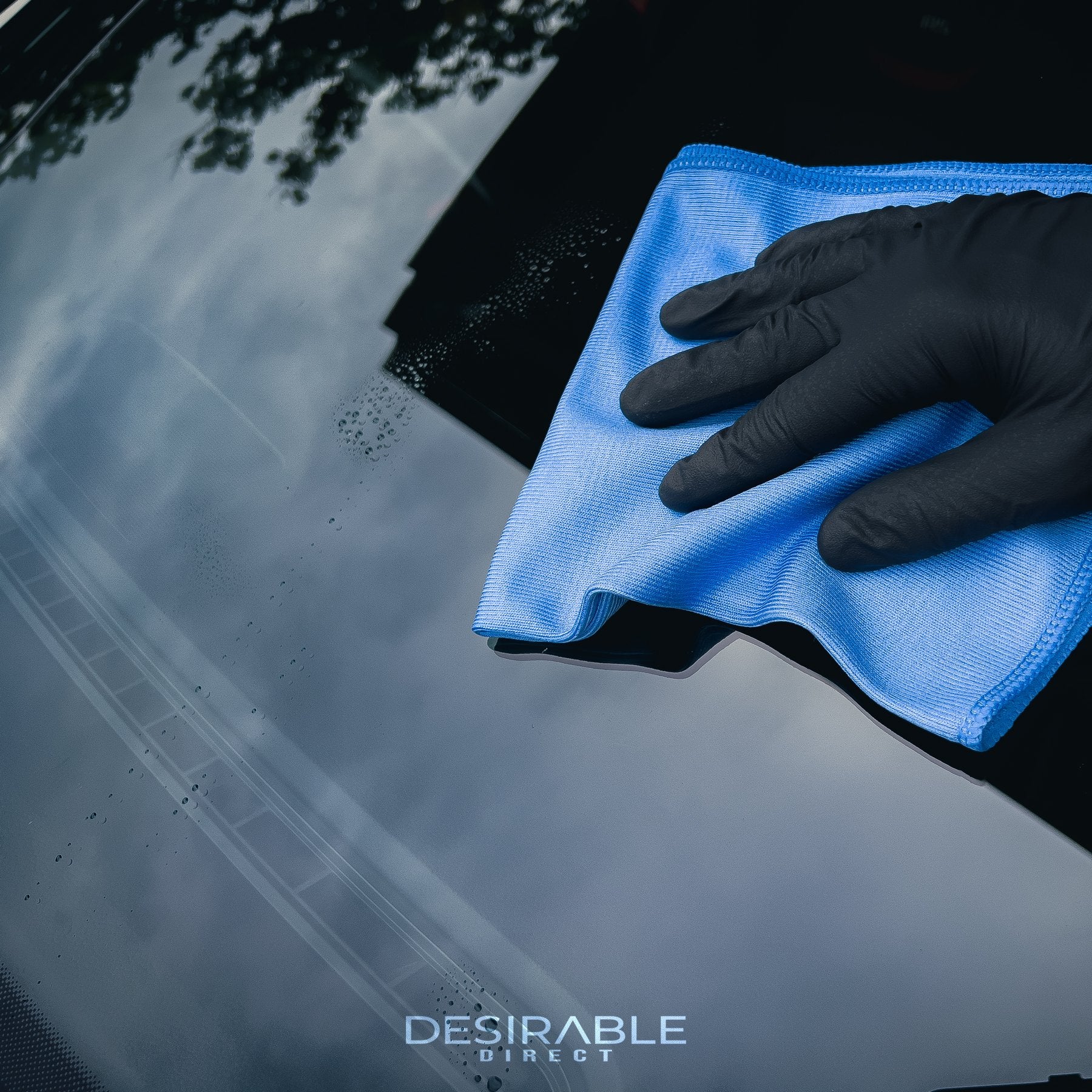 Car care microfibre blue glass cloth 40x40cm cleaning a car windscreen using car window cleaner. The person holding the cloth is wearing black gloves.