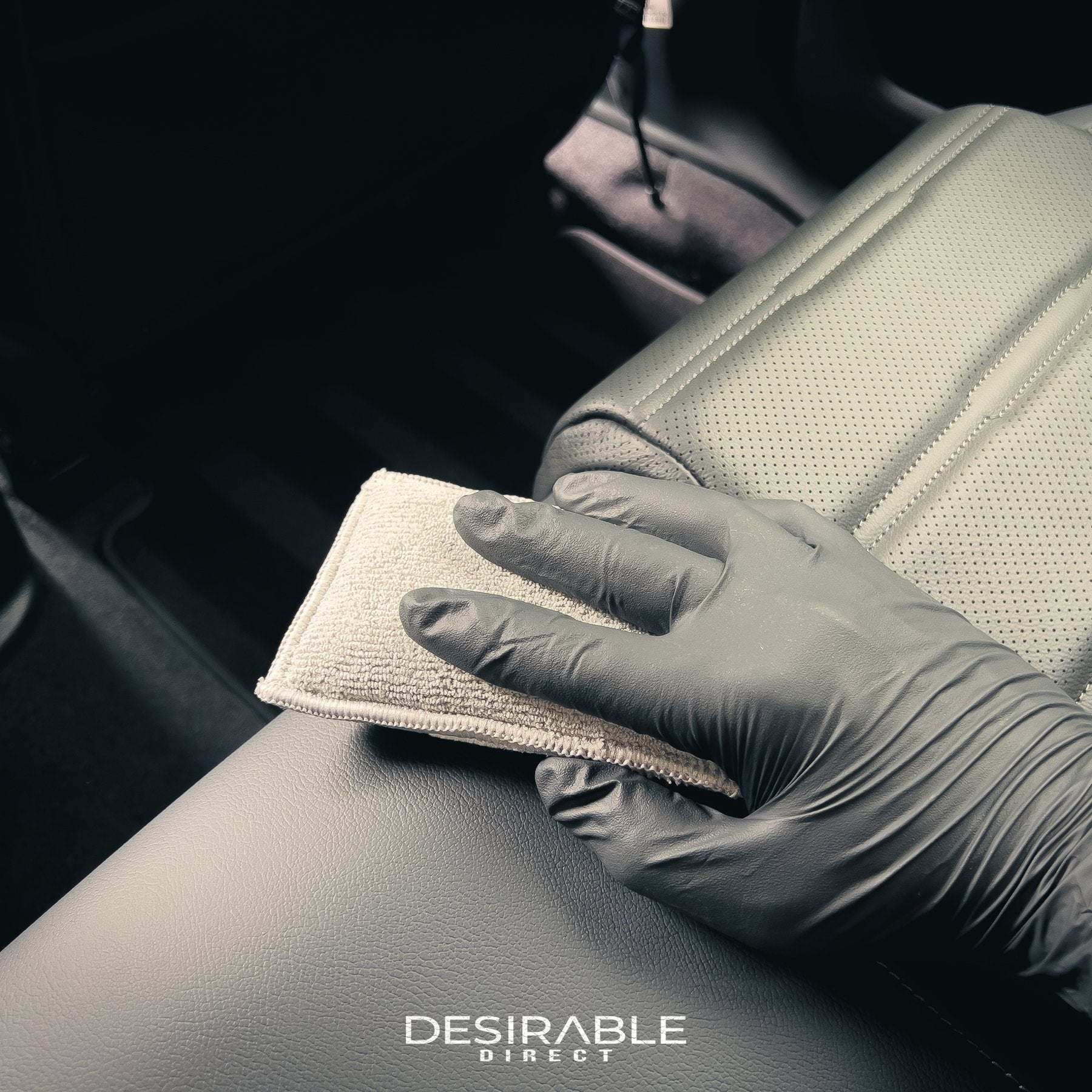 Car care one microfibre grey interior scrub pad cleaning the car seats at the back of the car. Somebody is holding the pad while wearing black gloves.