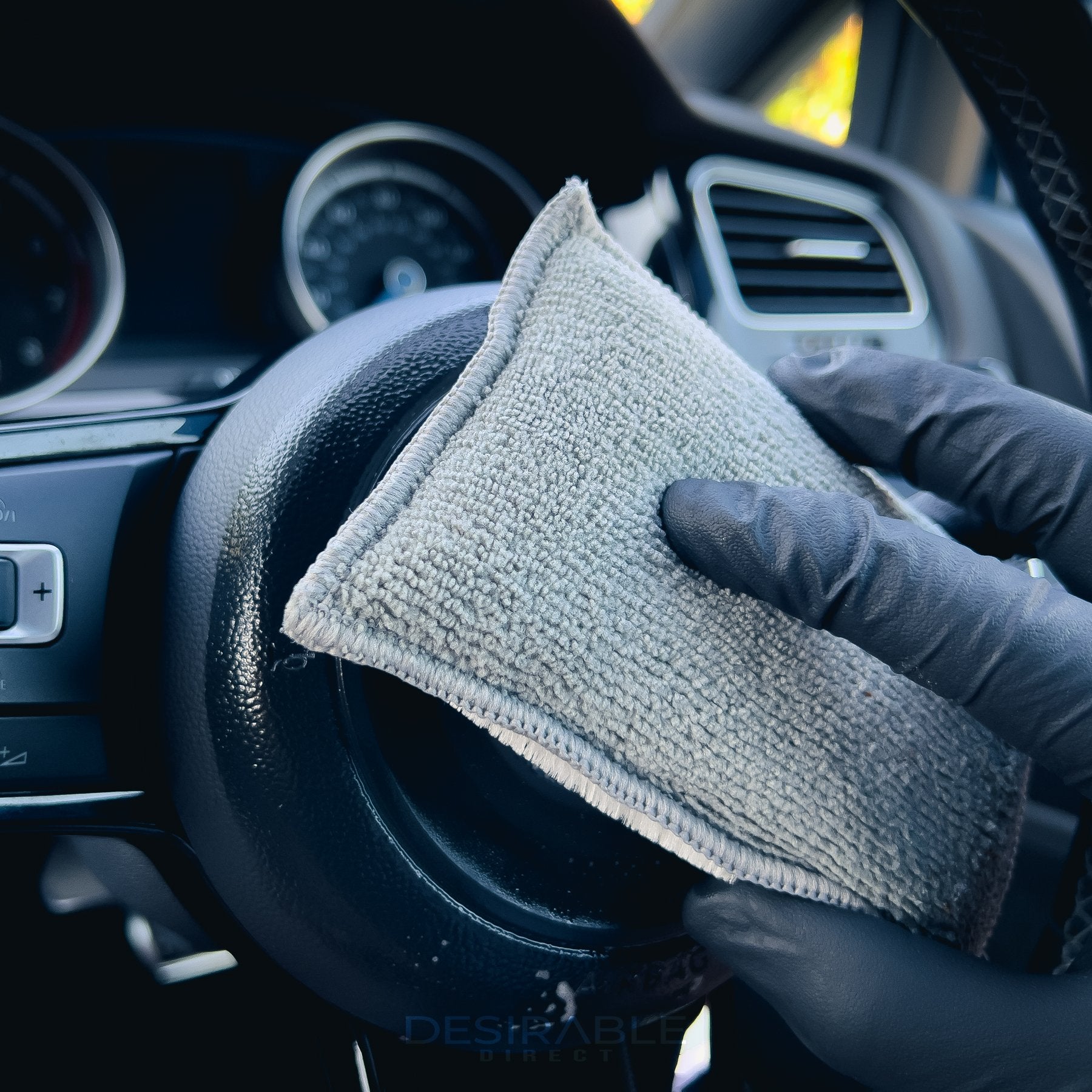 Car care one microfibre grey interior scrub pad cleaning the steering wheel of a volkswagen golf r. Somebody is holding the pad while wearing black gloves.