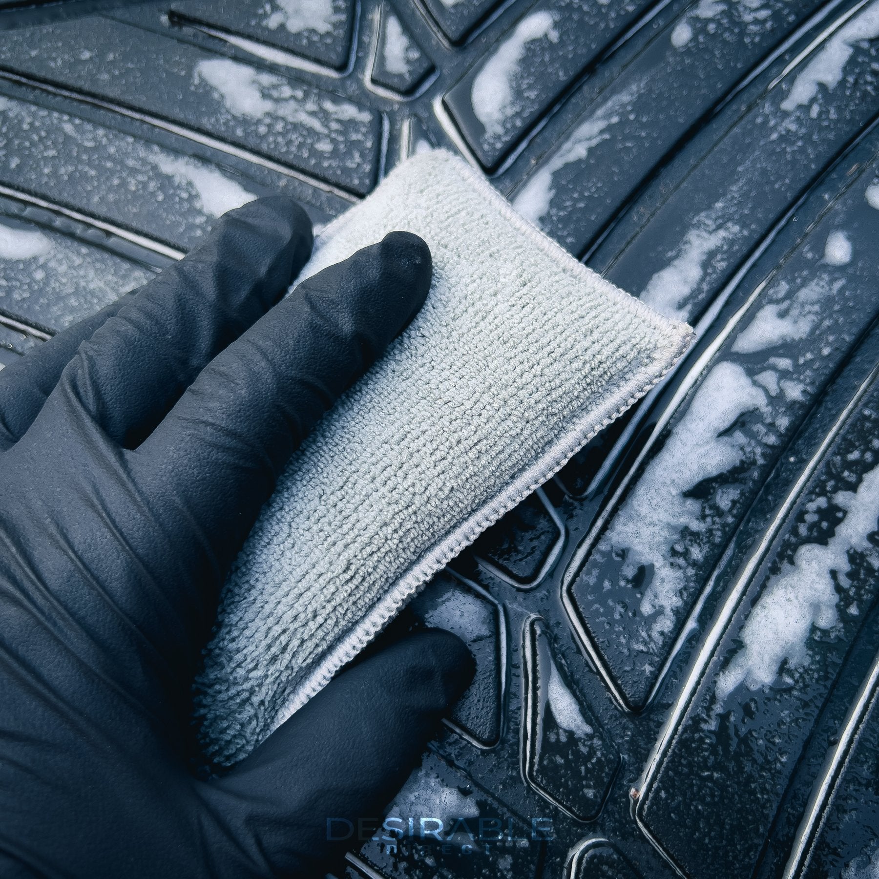 Car care one microfibre grey interior scrub pad cleaning a rubber car floor matt. Somebody is holding the pad while wearing black gloves.