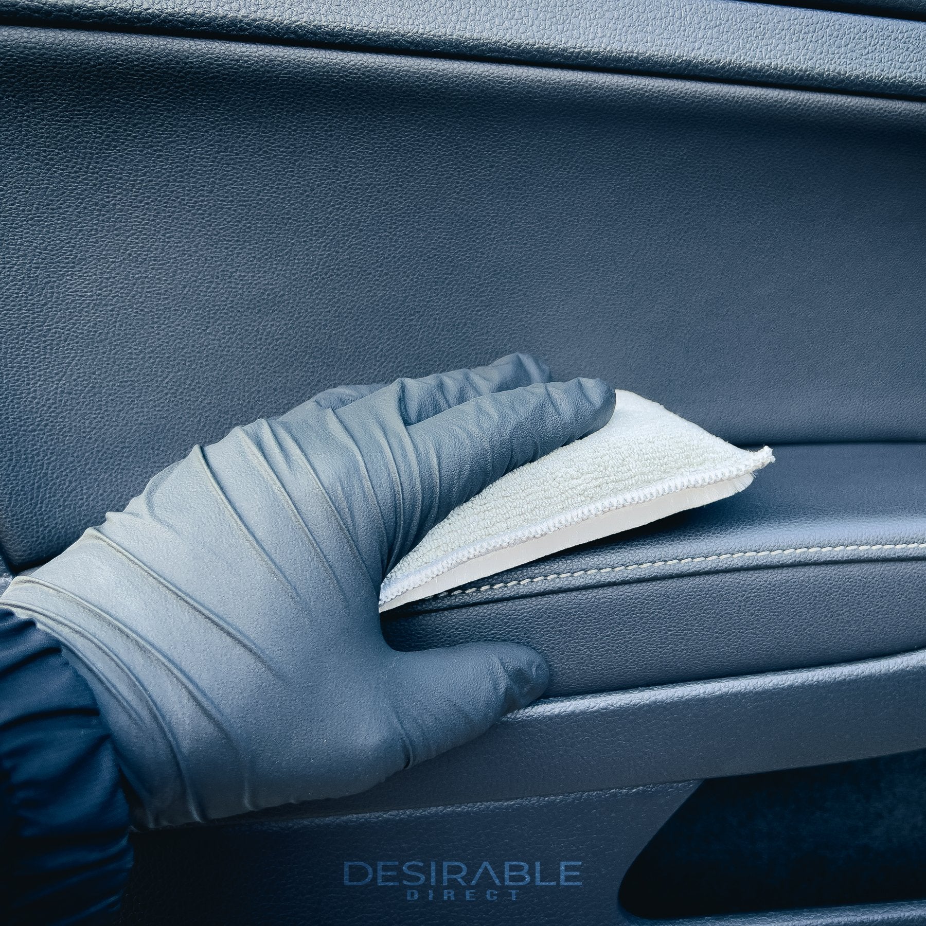 Car care one microfibre grey interior scrub pad is cleaning the leather passenger side door armrest of a car. Somebody is holding the pad and wearing black gloves.