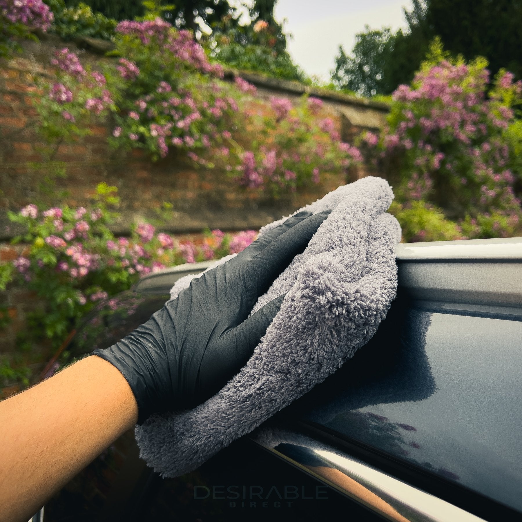 Car care plush grey microfibre cloth cleaning the roof of a grey car with pink flowers in the background.