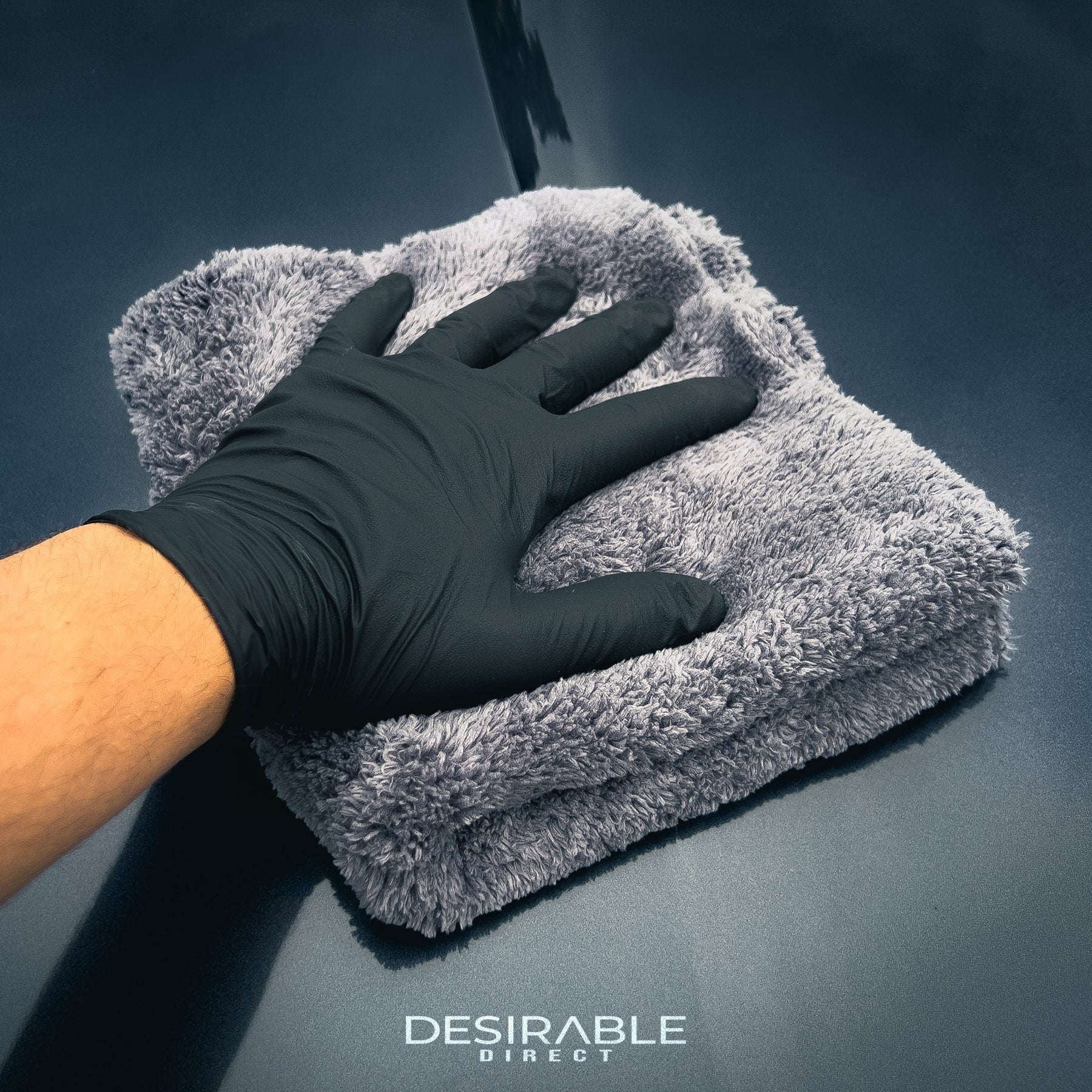 Car care plush grey microfibre cloth cleaning the bonnet of a grey car. The person holding the cloth is wearing black gloves.