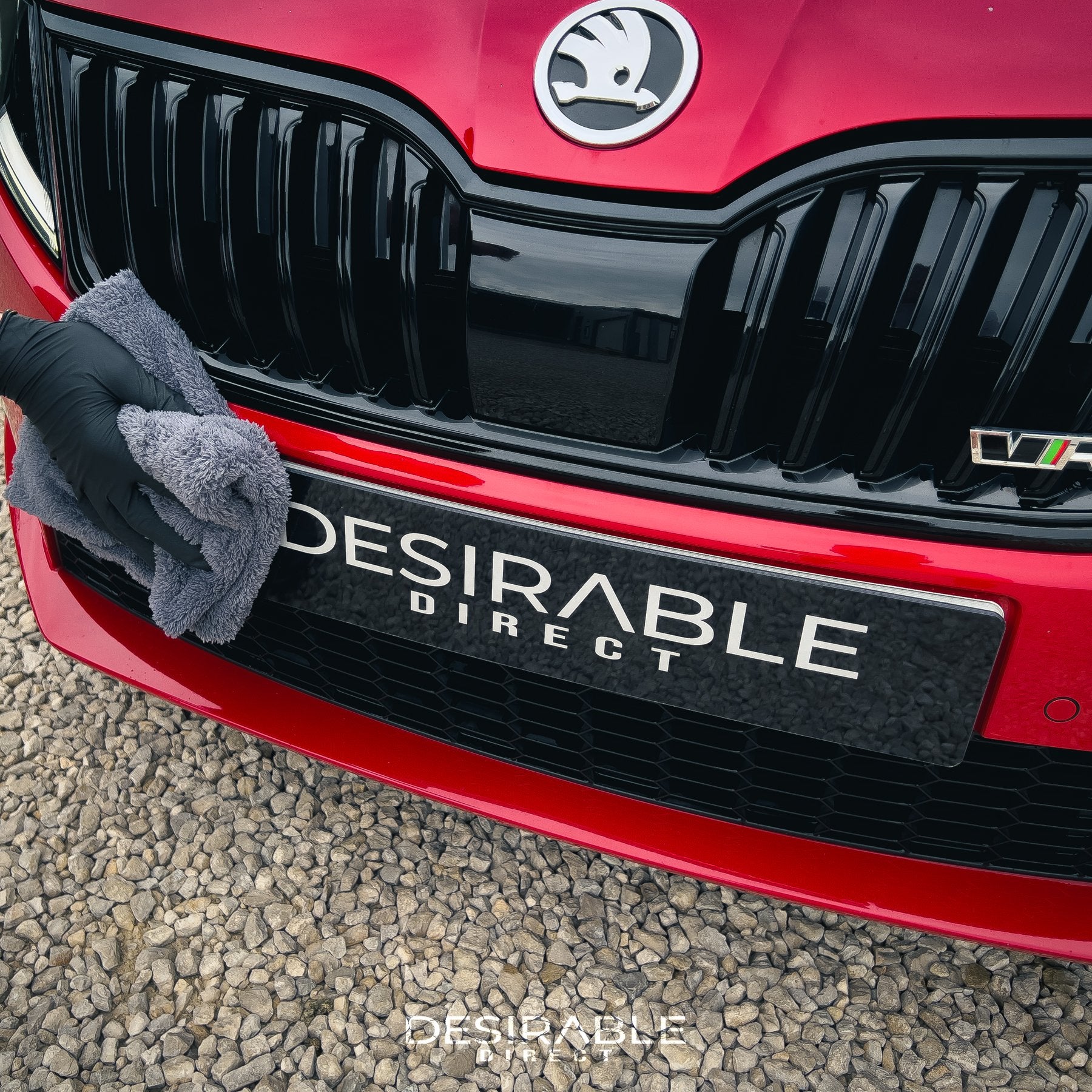 Car care plush grey microfibre cloth cleaning the front of a red car. The person holding the cloth is wearing black gloves.