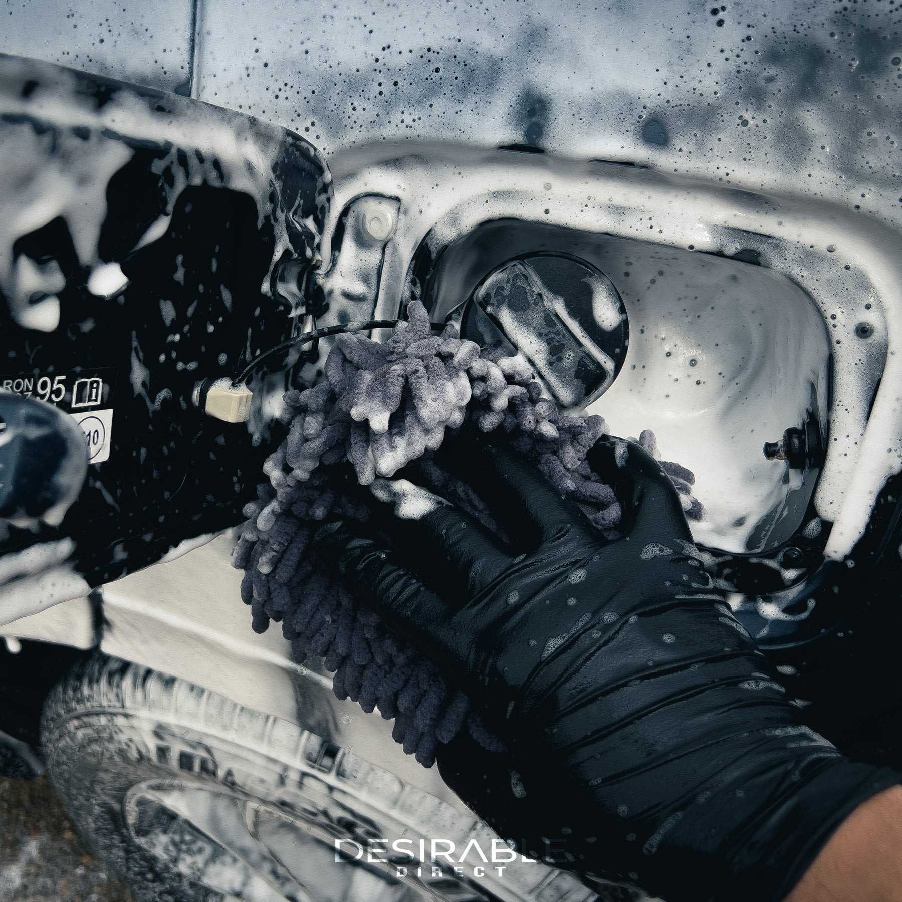 Car care chenille grey wheel and trim noodle wash pad cleaning the fuel filler cap of a car. The car is covered in car shampoo and the person holding the mitt is wearing black gloves.
