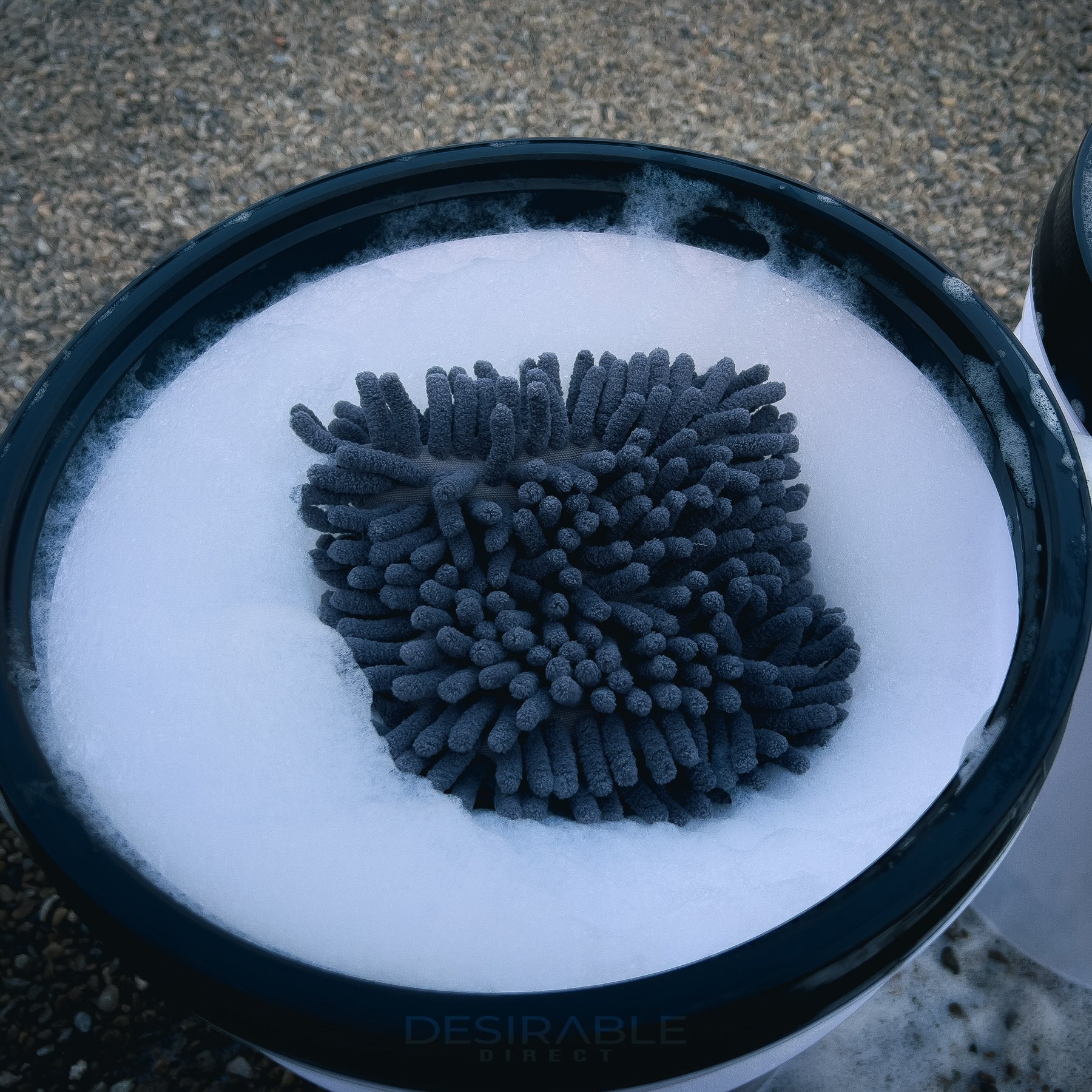 Car care chenille grey wheel and trim noodle wash pad submerged in a bucket of water filled with car shampoo.
