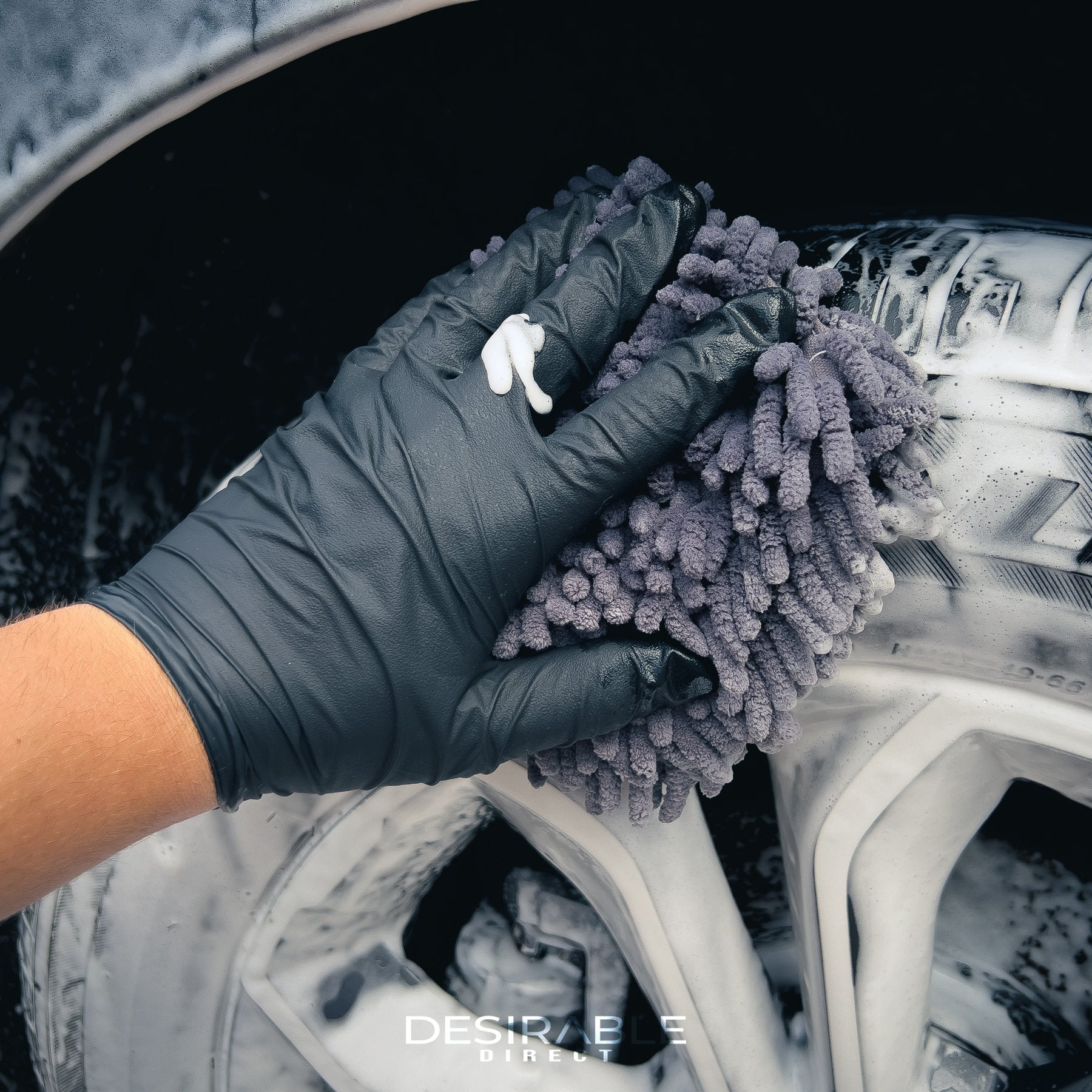 Car care noodle grey small pad cleaning the tyre of as grey car covered in car shampoo. The person holding the mitt is wearing a black glove.
