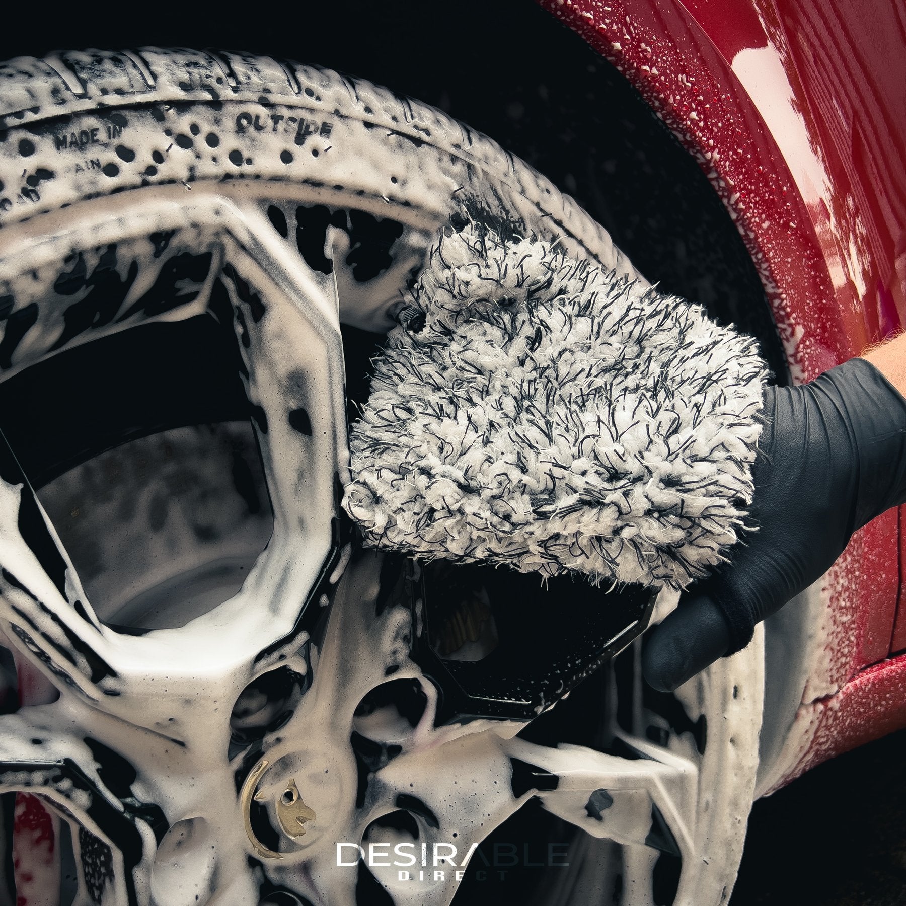Car care microfibre black and white wheel and trim wash mitt cleaning the wheels of a red car with black wheels. The wheels are covered in car shampoo. This is a close-up image to show the mitt.