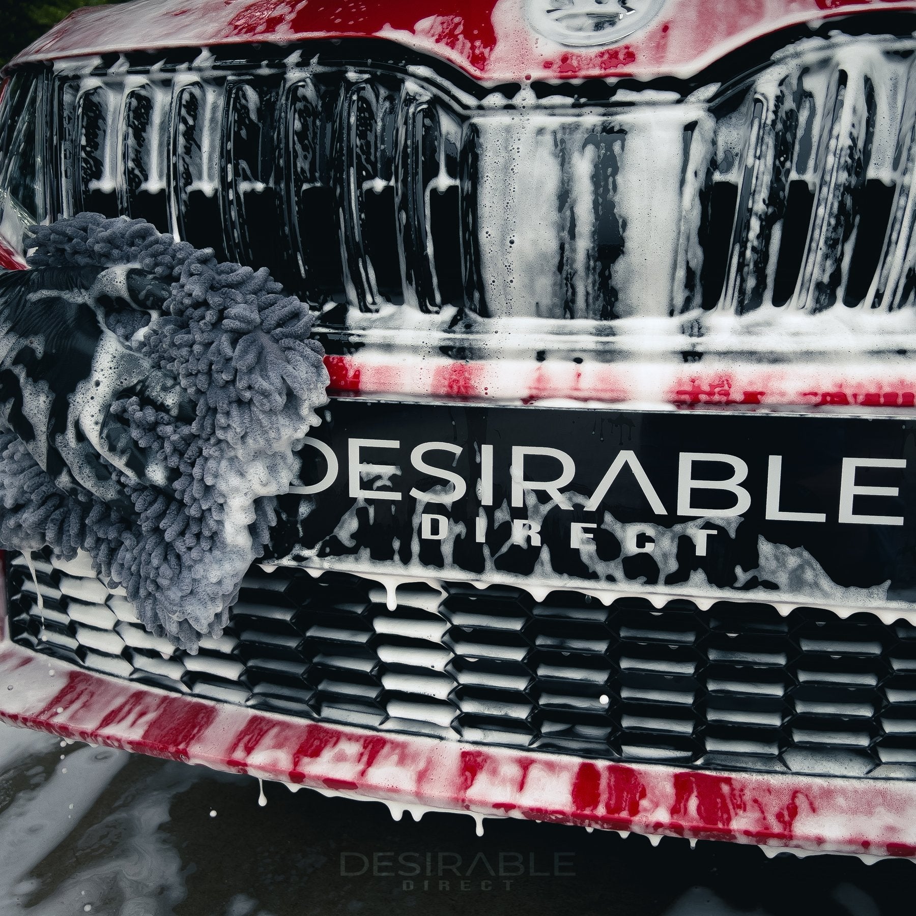 Car care noodle grey large pad cleaning the front of a red car. The person holding the pad is wearing black gloves.