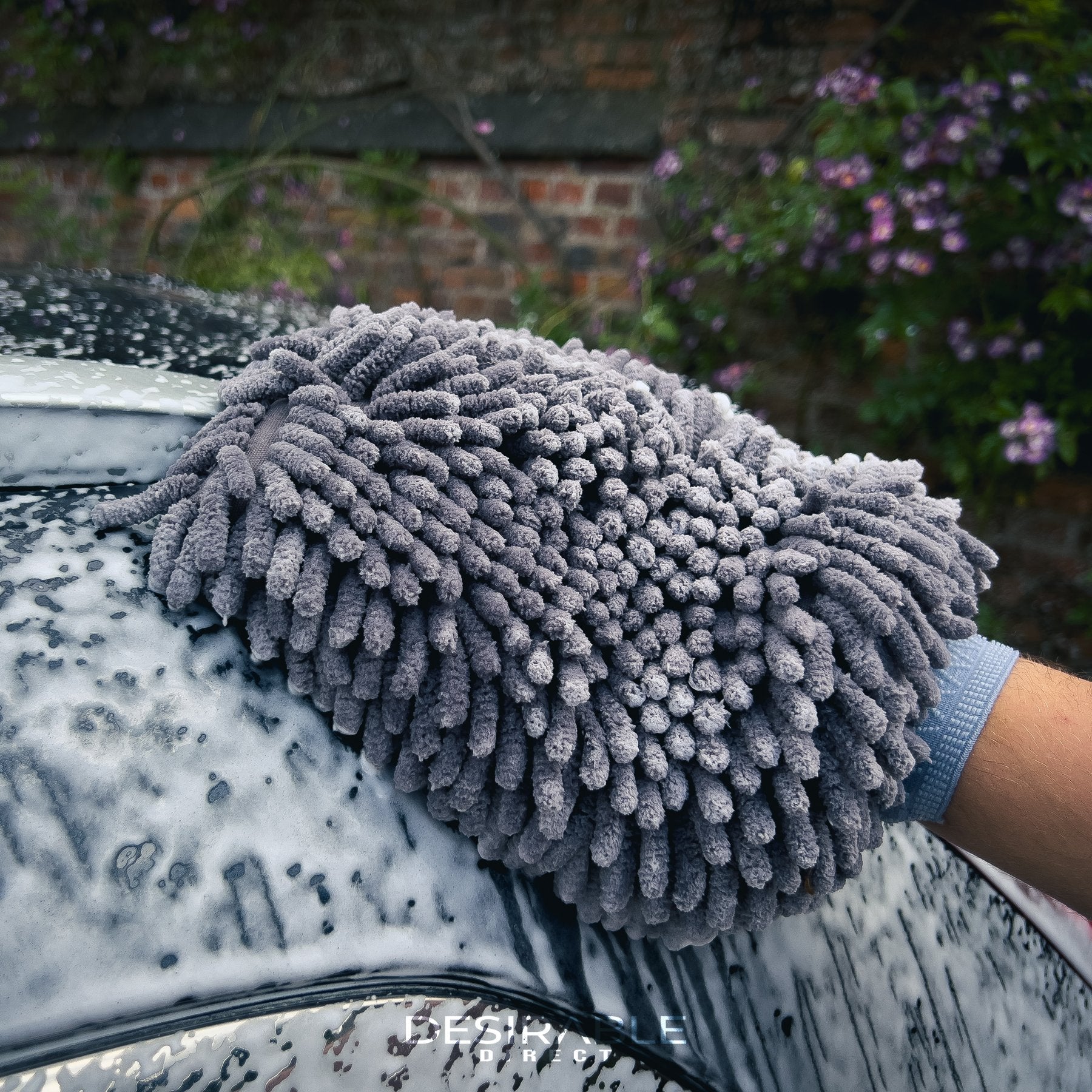 Car care noodle chenille grey wash mitt cleaning the top section of a grey car covered in car shampoo. With a wall and pink flowers in the background.