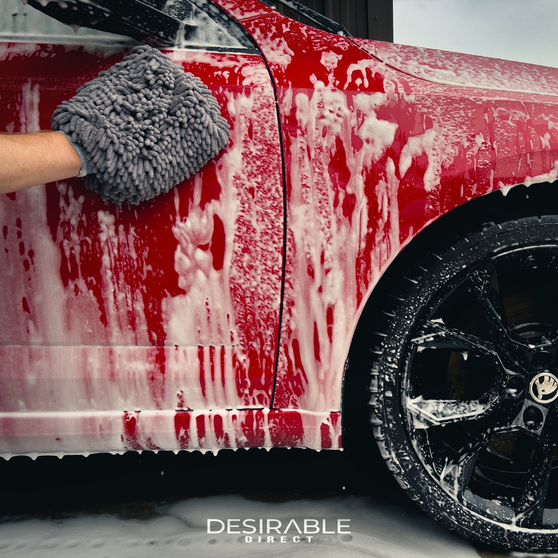 Car care noodle chenille grey wash mitt cleaning the side of a red car with black wheels covered in car shampoo.