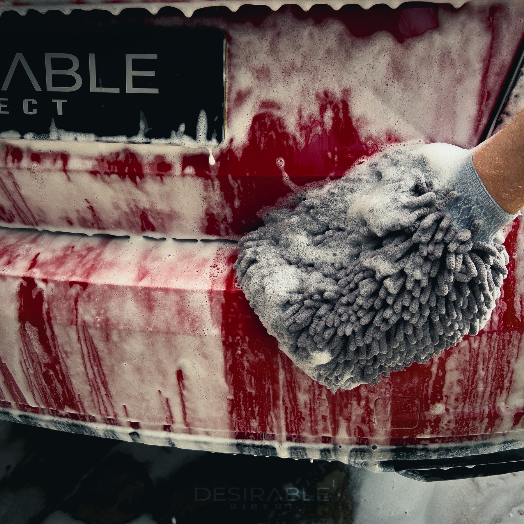 Car care noodle chenille grey wash mitt cleaning the back of a red car with black wheels covered in car shampoo.