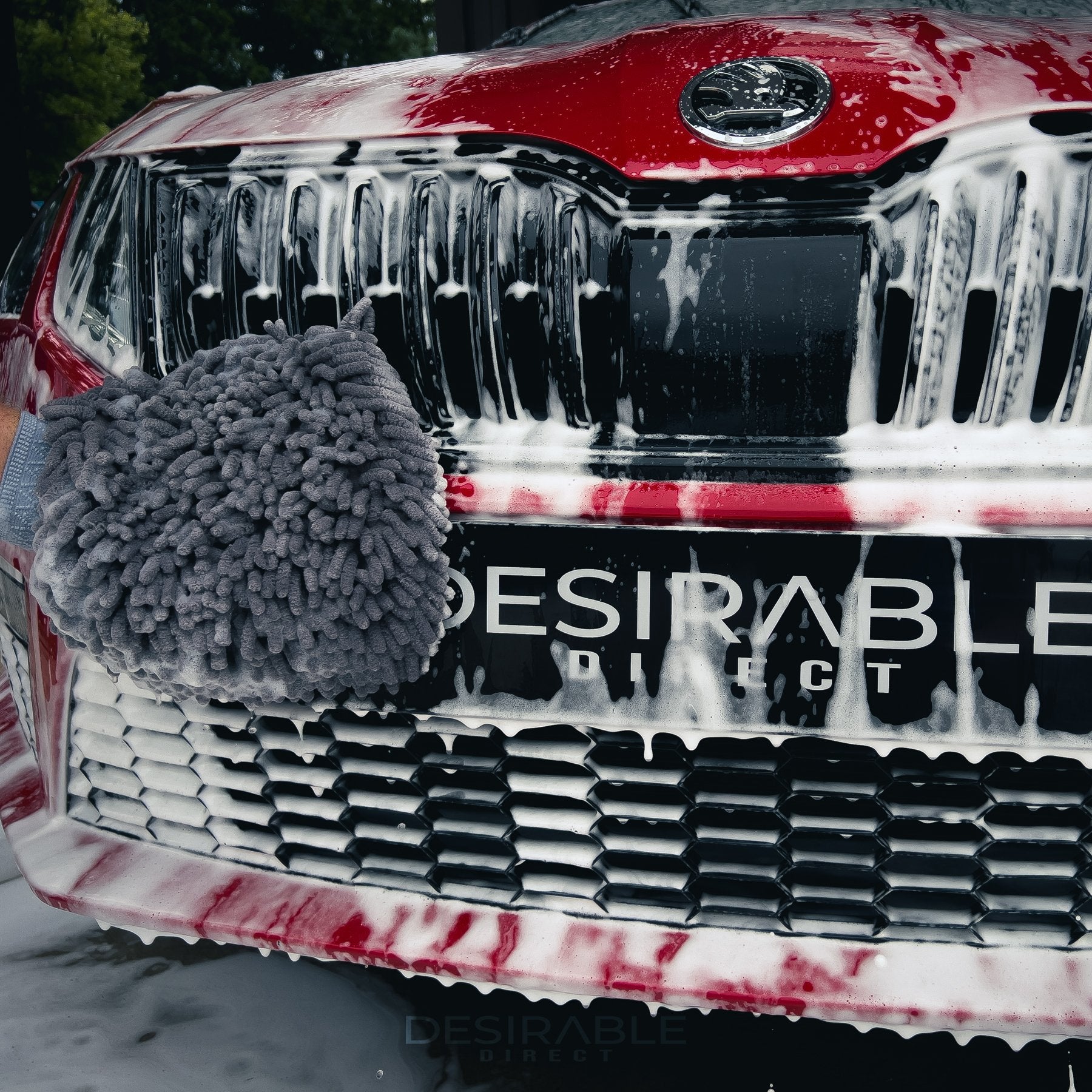 Car care noodle chenille grey wash mitt cleaning the front of a red car covered in car shampoo. The registration plate is covered by a black branded plate with desirable direct printed on the front.