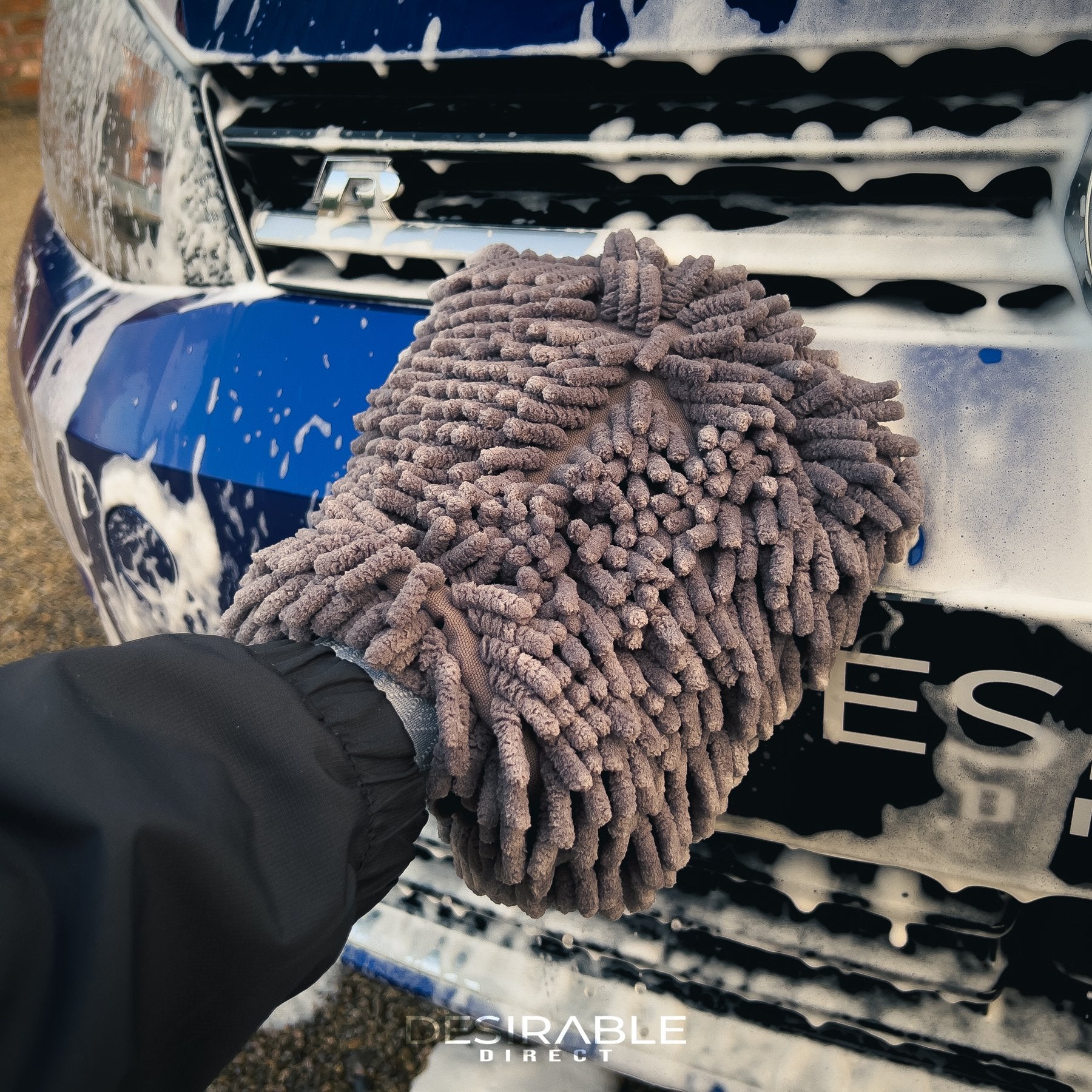 Car care noodle chenille grey wash mitt cleaning the front of a blue volkswagen golf r covered in car shampoo.