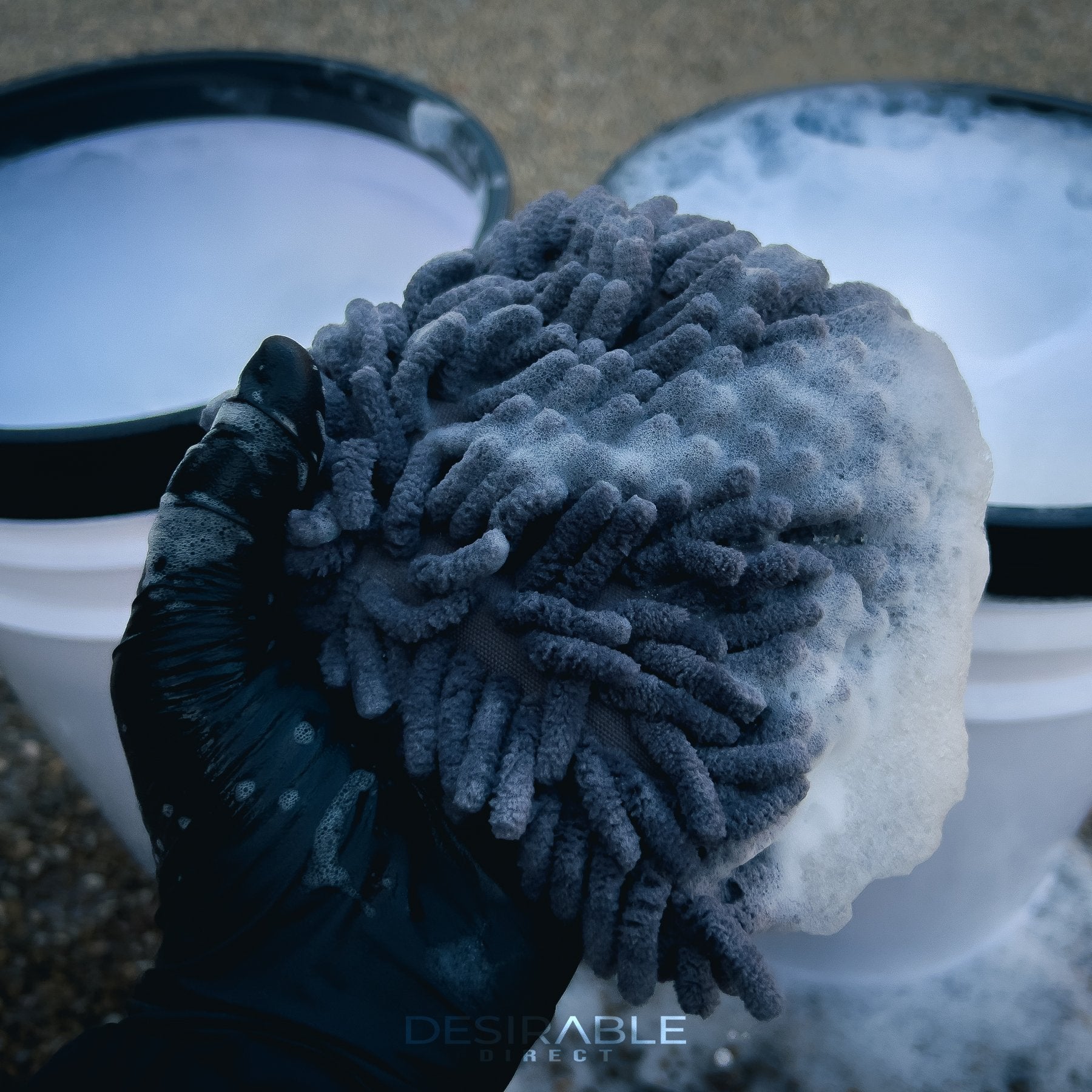 Car care noodle grey small pad covered in car shampoo with two car cleaning buckets in the background filled with water and car shampoo.